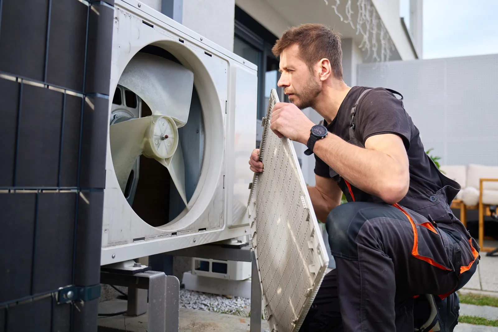Technician kneeling to remove the front grille of an outdoor heat pump, exposing the fan