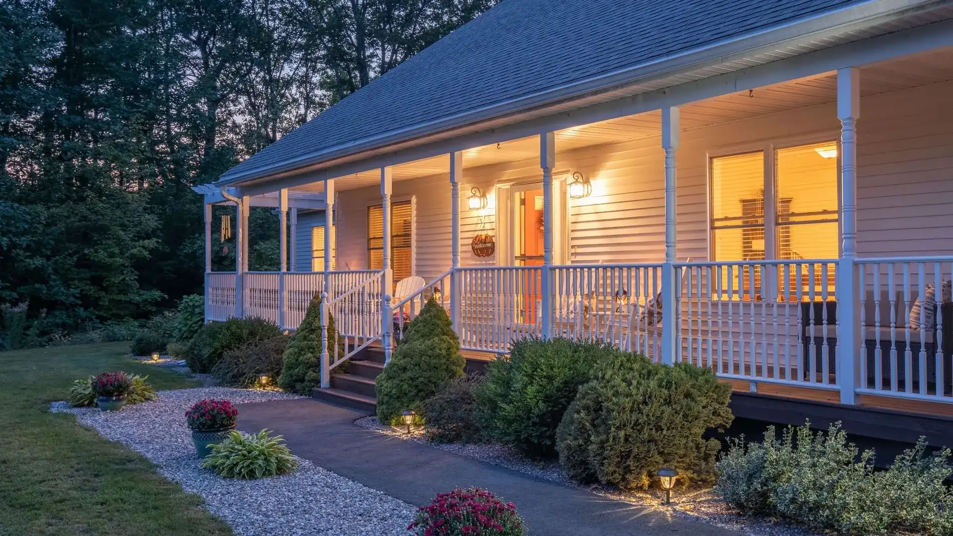 Single-story house with a long lit front porch and landscaped beds at dusk