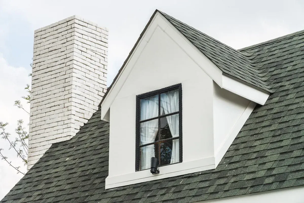 White dormer window set into a green-grey shingle roof beside a white brick chimney