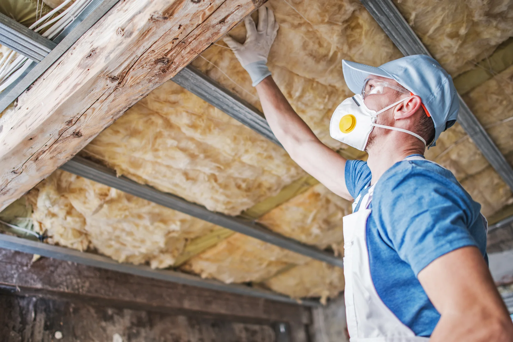 Worker in a dust mask and cap checking yellow insulation between old attic rafters