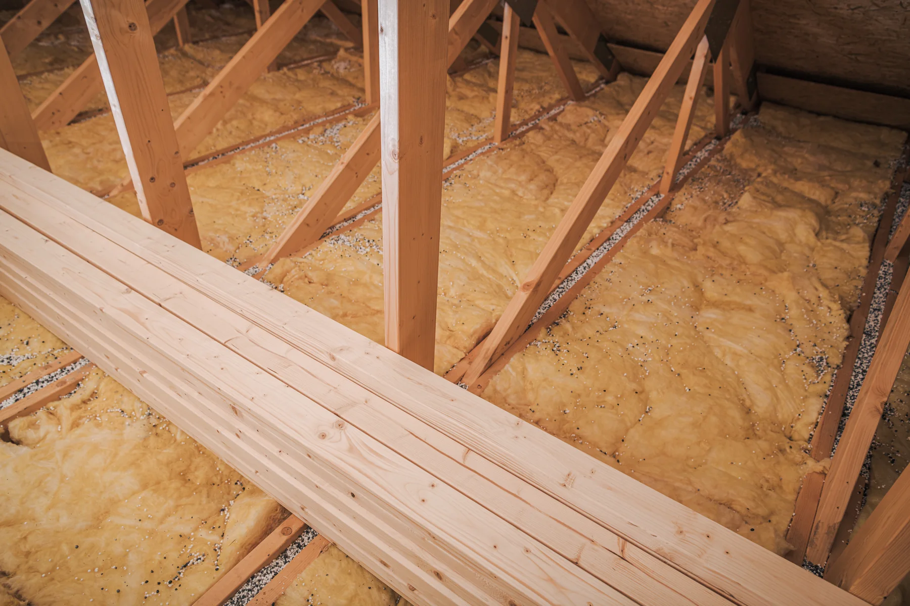 Attic floor covered with yellow fiberglass insulation between wooden roof trusses and stacked lumber