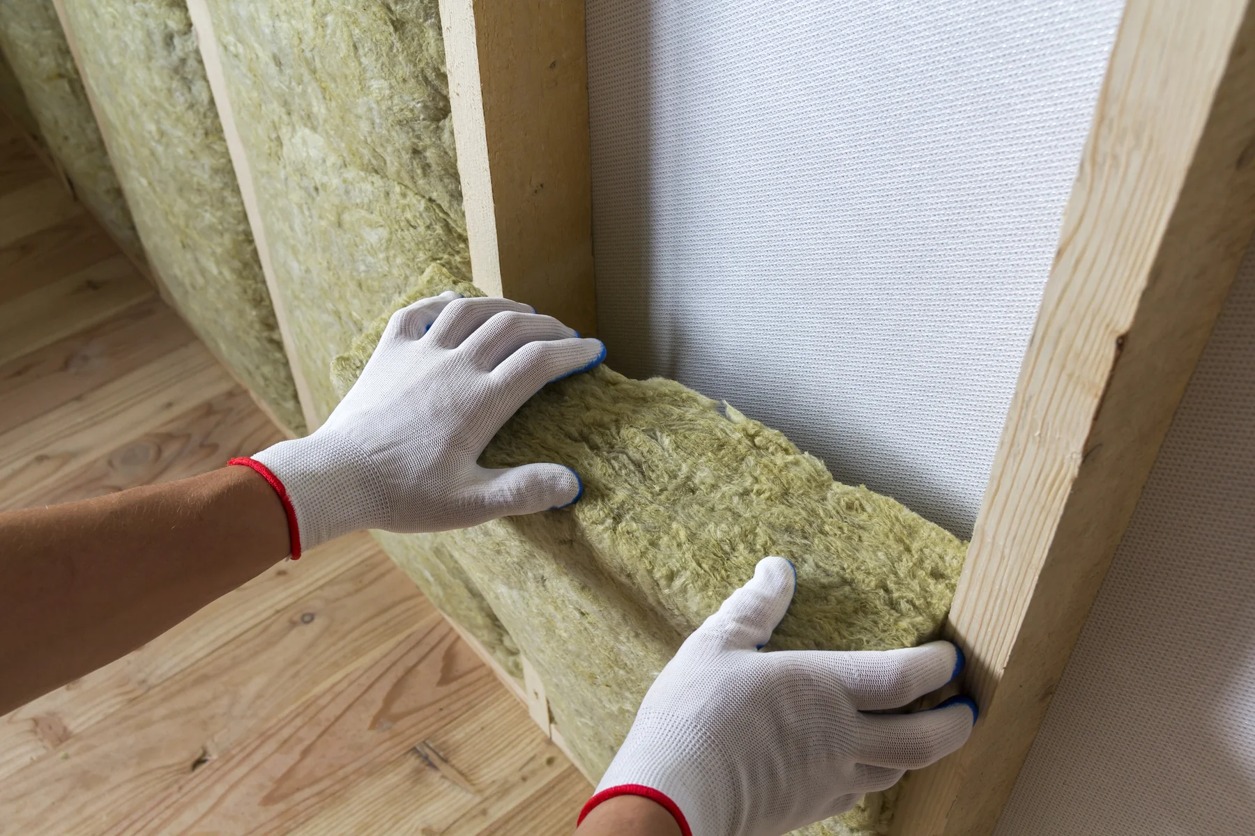 Gloved hands pressing a mineral wool insulation batt between wooden wall studs