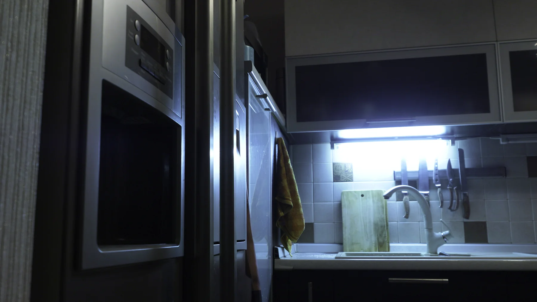 Dark kitchen at night with one under-cabinet light glowing near the refrigerator and oven