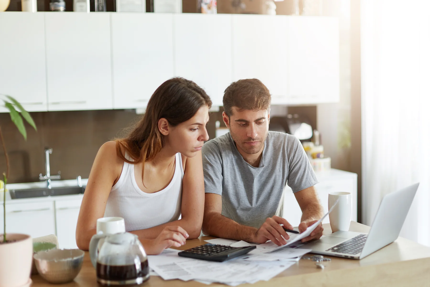 Young couple at a kitchen table reviewing paperwork with a calculator and laptop