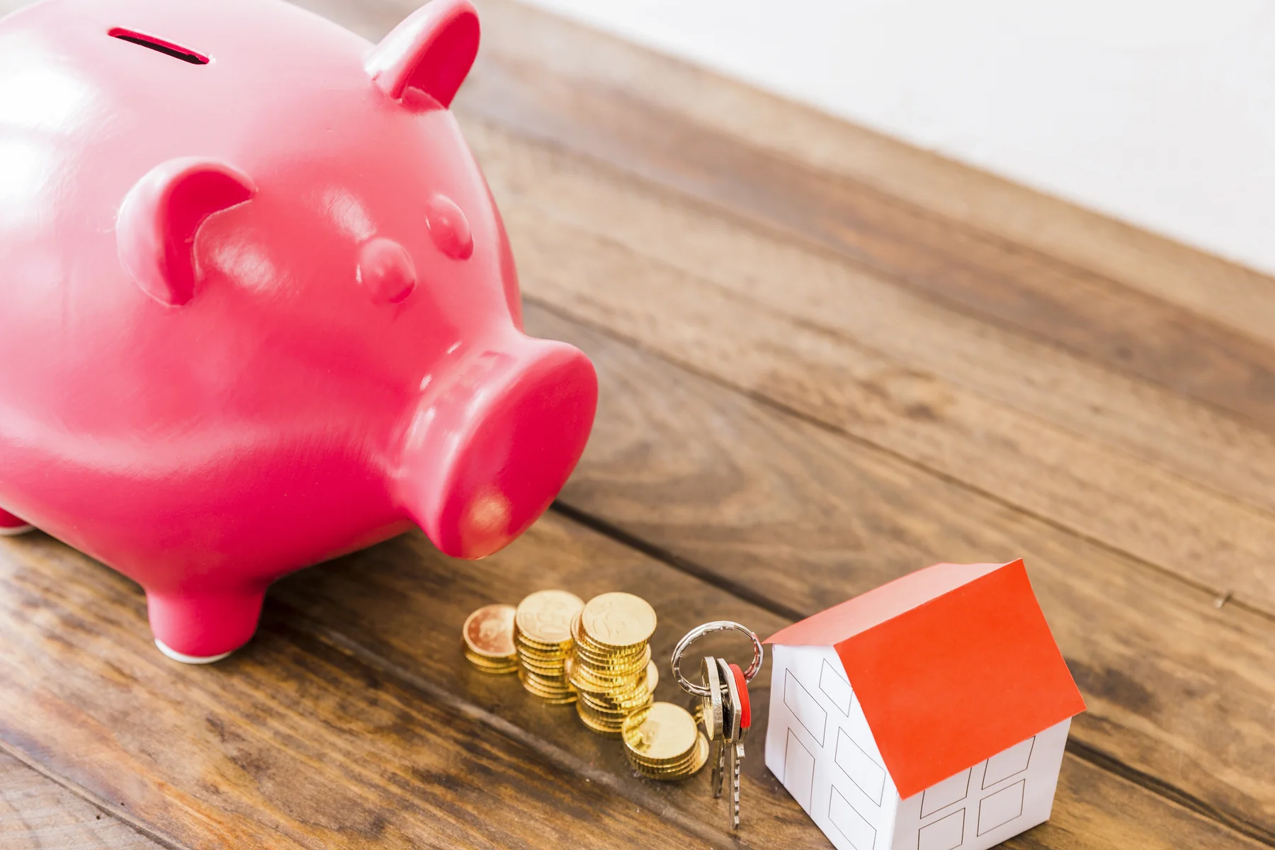 Pink piggy bank, stacked coins, house keys and a small paper house on a wooden desk