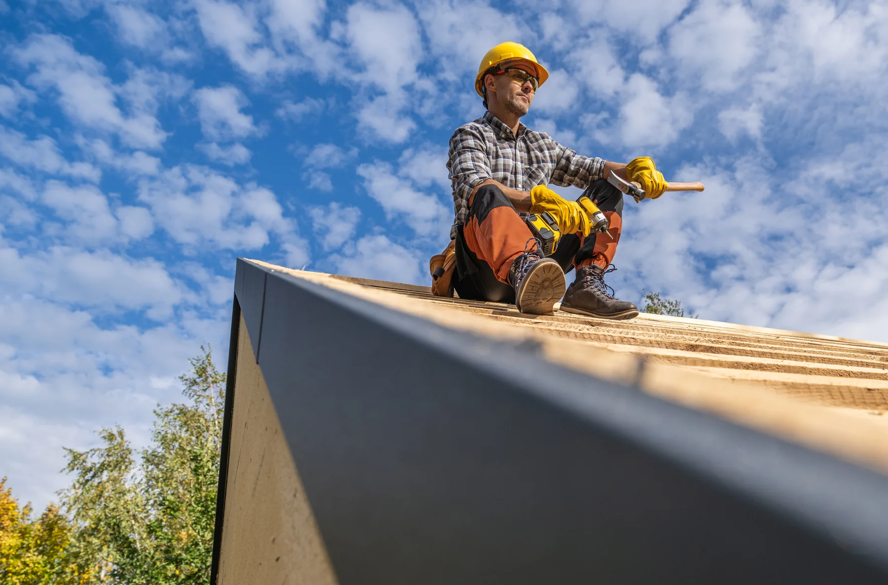 Roofer in a hard hat and gloves sitting on bare roof decking under a blue sky