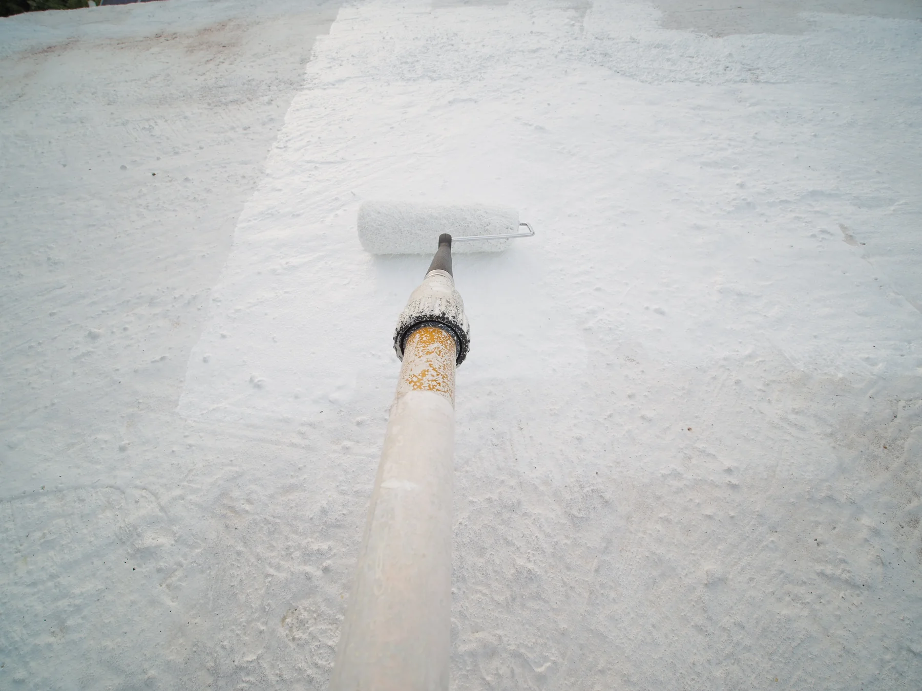 Paint roller on a long pole spreading white reflective coating across a flat roof surface