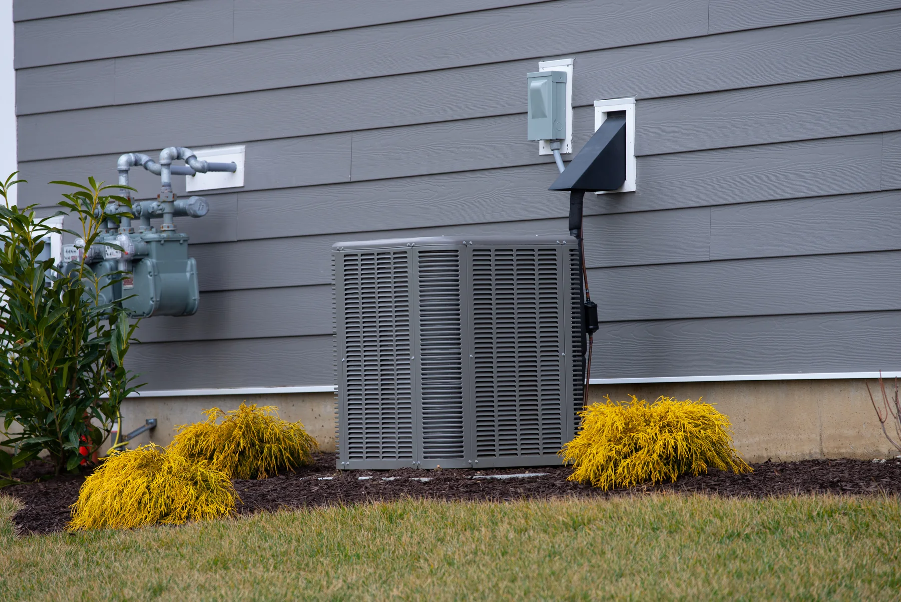 Outdoor central air conditioner condenser beside gray siding with a gas meter and shrubs