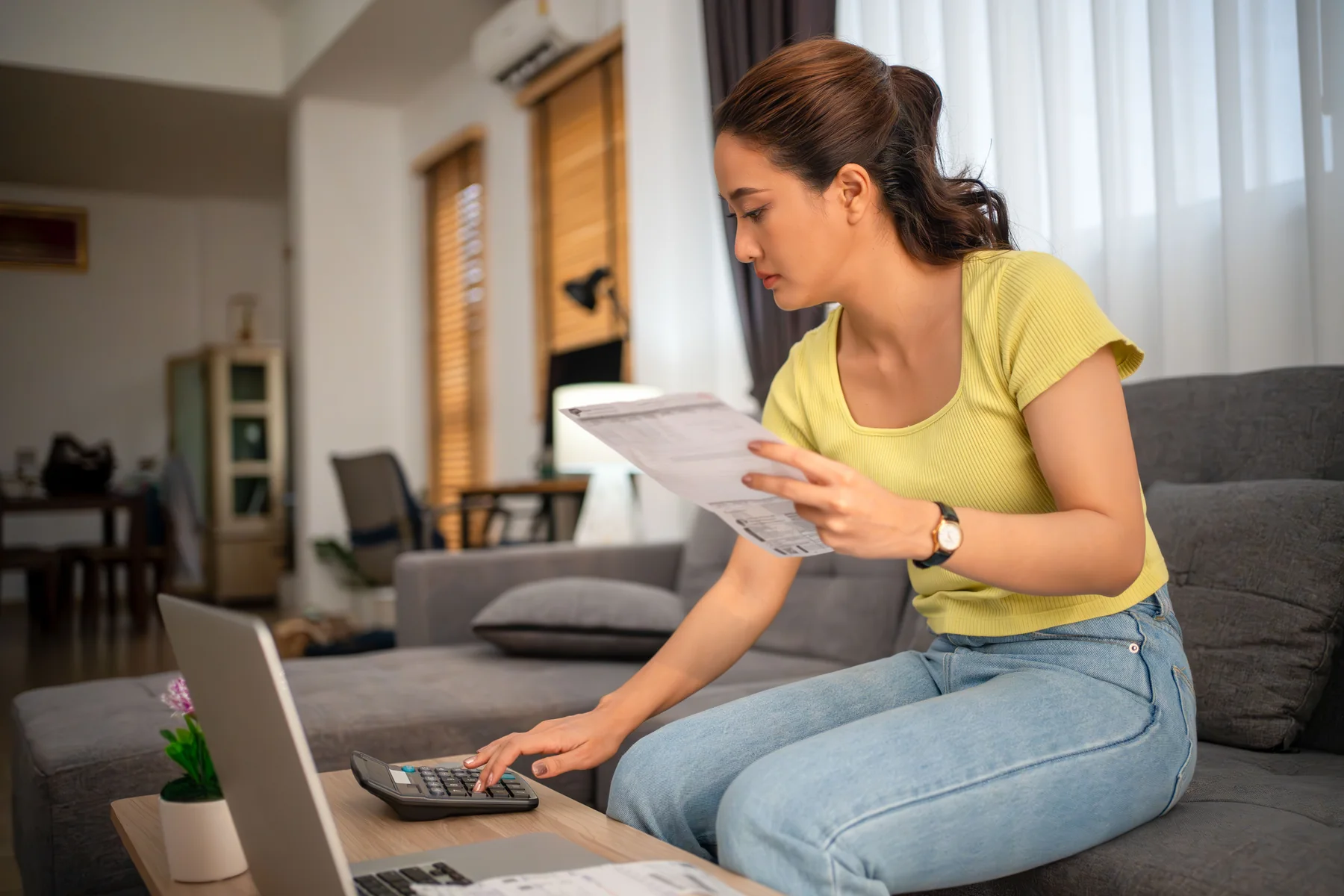 Woman on a sofa holding a household bill while tapping numbers into a calculator