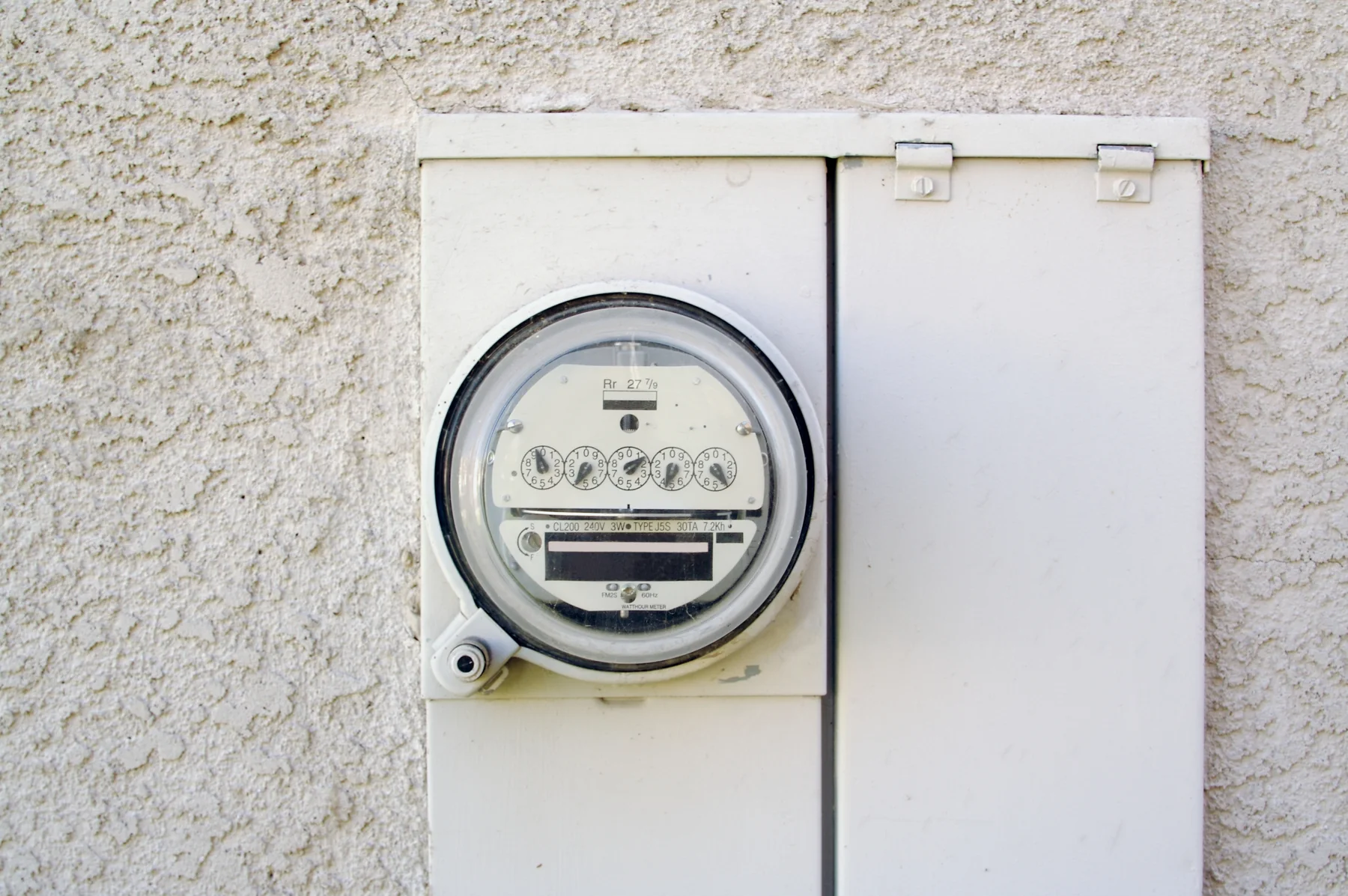 Round electric meter with dial registers and a small digital display on a stucco wall