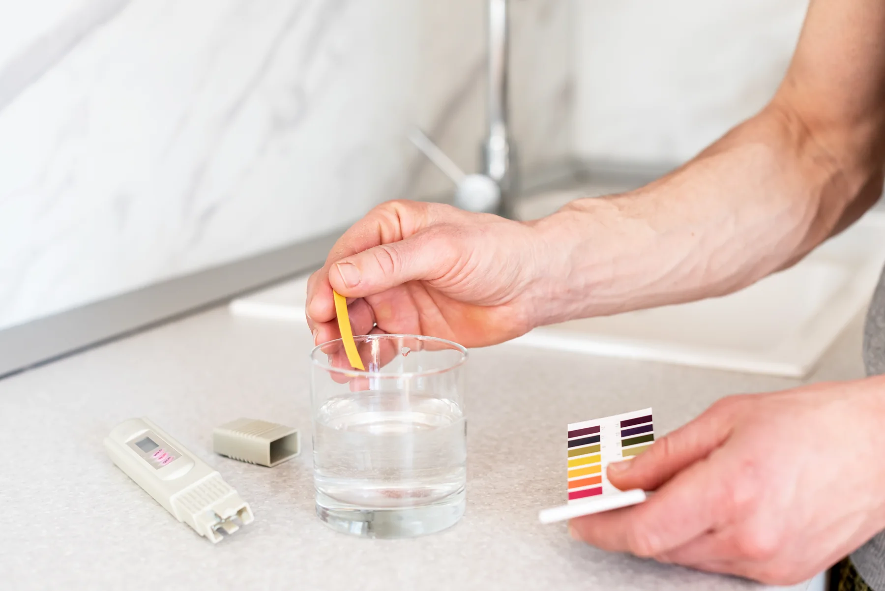 Hand dipping a pH test strip into a glass of tap water beside a TDS meter